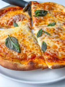 A close-up of an Easy Homemade Pizza topped with fresh basil leaves, cut into slices on a white plate with a pizza cutter nearby.