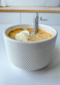 A white textured cup filled with coffee and a scoop of vanilla ice cream—a classic affogato recipe—finished with an ornate silver spoon. The cup sits on a white surface, the kitchen background softly blurred.