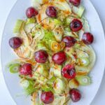 A white oval plate with a fresh salad of sliced fennel, celery, and whole cherries, drizzled with a brown vinaigrette and garnished with celery leaves and fennel fronds—a refreshing contrast to a Peaches and Cream Dessert.