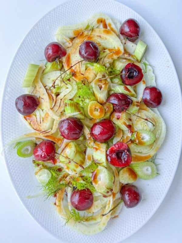 A white oval plate with a fresh salad of sliced fennel, celery, and whole cherries, drizzled with a brown vinaigrette and garnished with celery leaves and fennel fronds—a refreshing contrast to a Peaches and Cream Dessert.