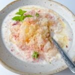 A bowl of yogurt topped with crushed ice, honey, and fresh mint leaves evokes a Peaches and Cream Dessert experience. A silver spoon rests in the bowl on a white surface, ready to enjoy this refreshing treat.