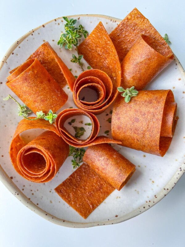 A plate with several rolled Fruit Leather (Lavashak) strips arranged in a floral pattern, garnished with small green herb sprigs on a white, speckled ceramic dish.