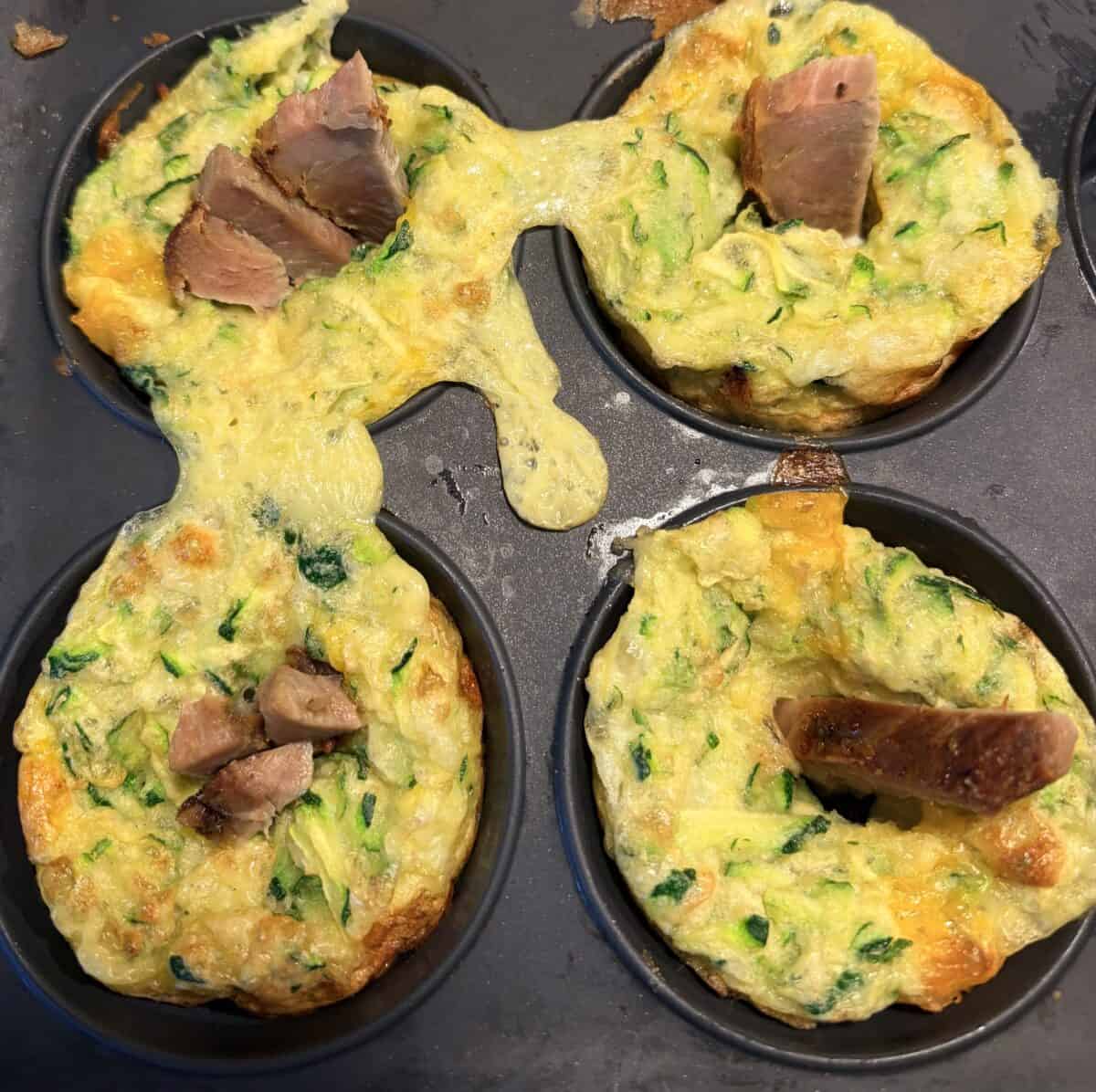 Four baked egg muffins, like a mini Zucchini Egg Bake, sit in a muffin tin topped with chopped meat. One muffin has overflowed and connected to another. Visible ingredients include cheese and green vegetables, possibly spinach or zucchini.