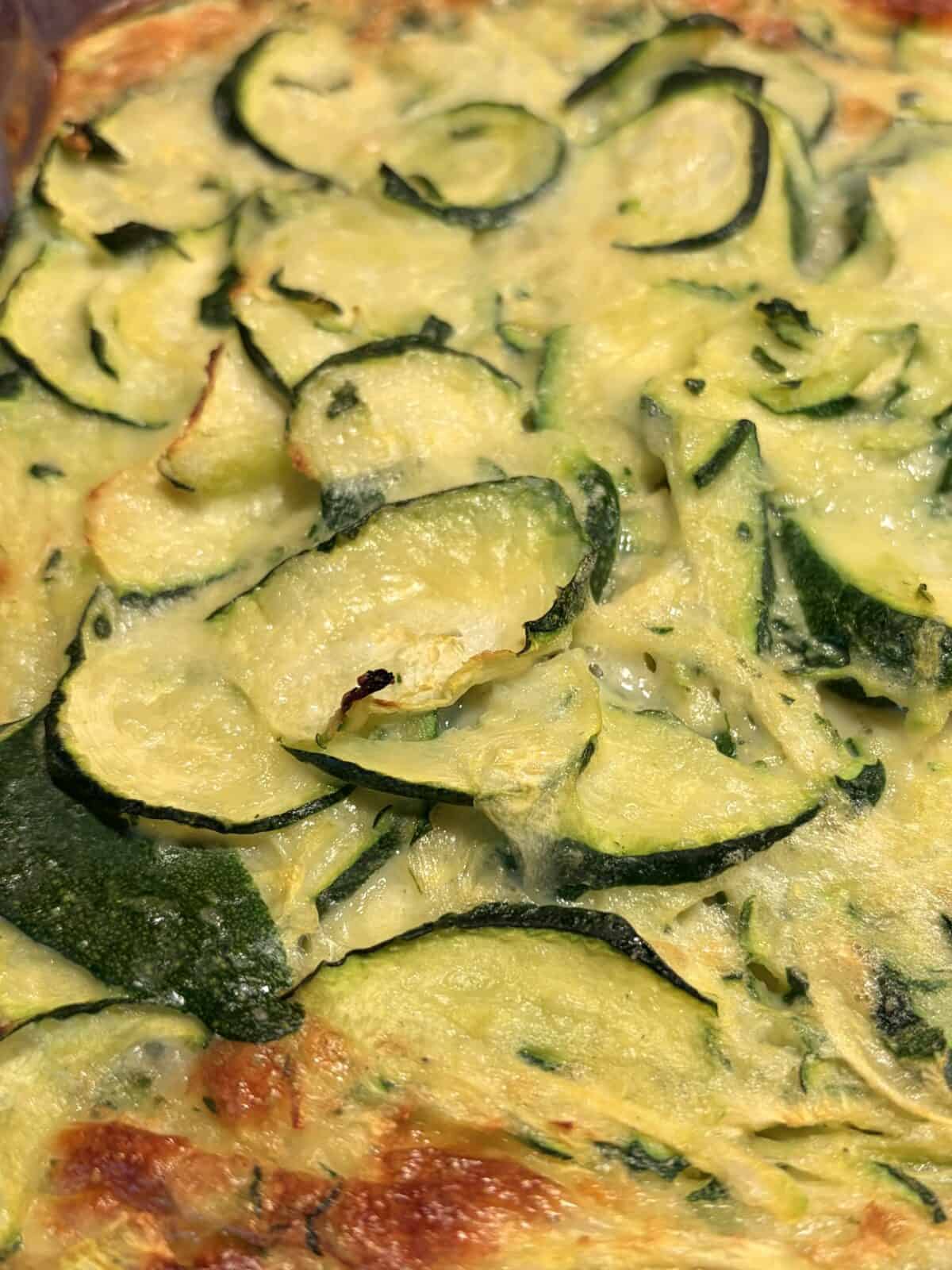 Close-up of a Zucchini Egg Bake featuring sliced zucchini coated in a creamy, cheesy mixture, with golden-brown edges and visible herbs throughout.