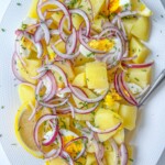 A white plate with Healthy Potato Salad featuring chopped boiled potatoes, sliced hard-boiled eggs, red onion rings, lemon wedges, and sprinkled fresh herbs. A fork rests on the plate.