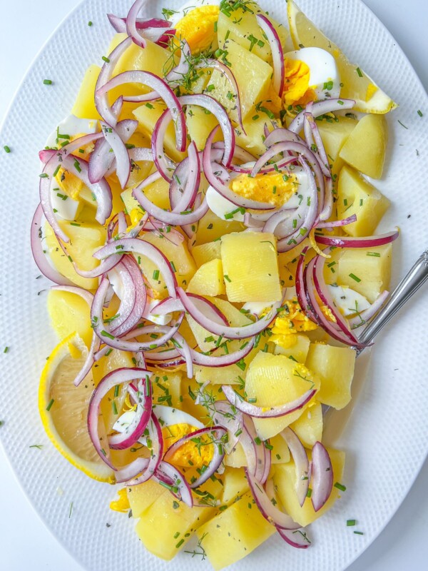 A white plate with Healthy Potato Salad featuring chopped boiled potatoes, sliced hard-boiled eggs, red onion rings, lemon wedges, and sprinkled fresh herbs. A fork rests on the plate.