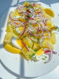 A white plate with sliced boiled potatoes, hard-boiled eggs, red onion, fresh herbs, and lemon wedges—ingredients often found in Olivier Salad—are all arranged under bright natural light.