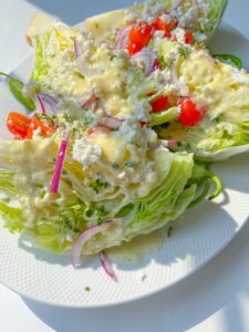 A classic Wedge Salad on a white plate, topped with cherry tomatoes, red onion slices, crumbled cheese, herbs, and creamy dressing, with sunlight highlighting the fresh ingredients.