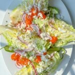 Three Wedge Salads are topped with cherry tomatoes, red onion slices, crumbled feta cheese, and chopped herbs, served on a white oval plate. The fresh and colorful Wedge Salad stands out under bright lighting.