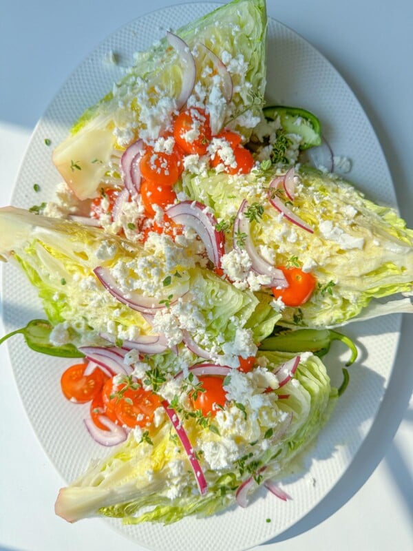 Three Wedge Salads are topped with cherry tomatoes, red onion slices, crumbled feta cheese, and chopped herbs, served on a white oval plate. The fresh and colorful Wedge Salad stands out under bright lighting.