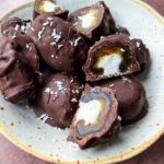 A ceramic plate filled with Bounty Dates—chocolate-covered treats, some whole and others cut open to reveal a white and caramel-colored filling—topped with flakes of sea salt.