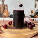 A glass of dark cherry juice sits on a wooden board, surrounded by fresh cherries. A single cherry garnishes the rim of the glass. The background shows a kitchen setting with utensils and cutting boards.