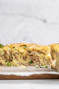 A savory slice of high protein breakfast casserole featuring fluffy cooked eggs and seasoned ground sausage, garnished with chopped fresh herbs, on a wooden board against a light background.