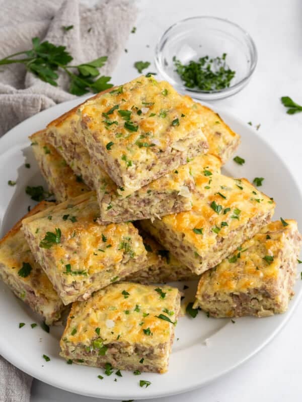 A plate of square-shaped sausage and cheese breakfast casserole pieces, garnished with chopped parsley. A small glass bowl with chopped herbs and a beige napkin are in the background.