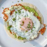 A fried egg with a runny yolk sits atop crispy cheese, mashed avocado, and a tortilla, garnished with chives—an irresistible twist inspired by the Viral Feta Fried Eggs trend.