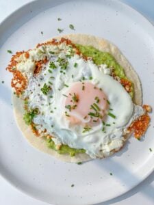 A fried egg with a runny yolk sits atop crispy cheese, mashed avocado, and a tortilla, garnished with chives—an irresistible twist inspired by the Viral Feta Fried Eggs trend.