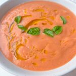 A bowl of creamy tomato soup, inspired by classic gazpacho, topped with a drizzle of olive oil and fresh basil leaves, served in a white bowl on a light background.
