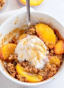 A bowl of Single Serve Peach Crisp topped with vanilla ice cream, sprinkled with cinnamon, and served with a spoon. This cozy dessert features baked peaches and a crumbly oat topping for the perfect treat.