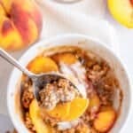 A close-up of a bowl filled with Single Serve Peach Crisp topped with creamy sauce. A spoon holds a bite of peaches and crisp. Fresh peaches, walnuts, and cinnamon sticks are nearby on a white surface.