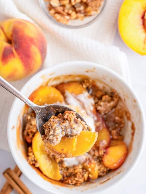 A close-up of a bowl filled with Single Serve Peach Crisp topped with creamy sauce. A spoon holds a bite of peaches and crisp. Fresh peaches, walnuts, and cinnamon sticks are nearby on a white surface.
