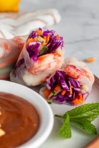 Fresh spring rolls filled with shrimp, purple cabbage, carrots, and cucumber are cut in half and displayed on a plate. They are accompanied by a small dish of peanut sauce and a sprig of mint. A striped cloth is in the background.