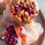 Two fresh spring rolls are placed on a plate. One is topped with a creamy peanut sauce, and both contain vibrant fillings of red cabbage, carrots, and cucumbers, wrapped in translucent rice paper.