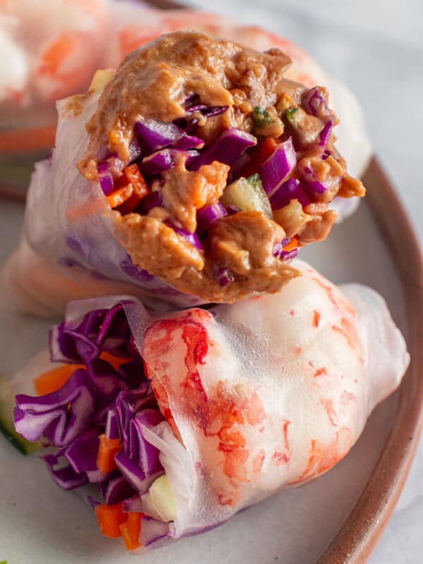 Two fresh spring rolls are placed on a plate. One is topped with a creamy peanut sauce, and both contain vibrant fillings of red cabbage, carrots, and cucumbers, wrapped in translucent rice paper.