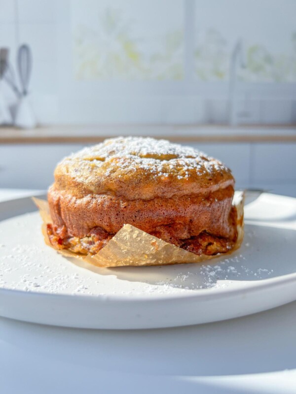 A close-up of a single serve banana bread, dusted with powdered sugar, sits on parchment paper atop a white plate, with a bright, blurred kitchen background.