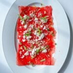 Watermelon Carpaccio: Thin slices of watermelon on an oval white plate, topped with crumbled white cheese, mint leaves, and a sprinkle of nuts or seeds, set against a white background.