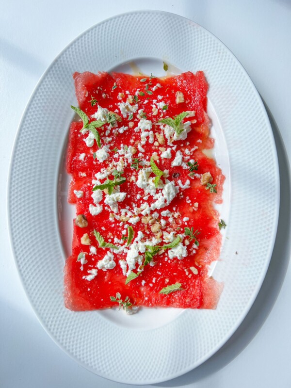 Watermelon Carpaccio: Thin slices of watermelon on an oval white plate, topped with crumbled white cheese, mint leaves, and a sprinkle of nuts or seeds, set against a white background.