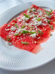 Thin slices of watermelon carpaccio are arranged on a white plate, topped with crumbled feta cheese, chopped nuts, and fresh green herbs. The dish is light and vibrant with a refreshing appearance.