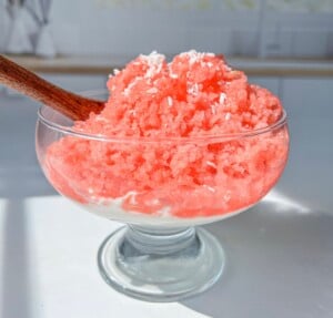 A glass dessert bowl filled with bright pink shaved ice topped with shredded coconut, sitting on a layer of white cream, with a wooden spoon sticking out from the side.
