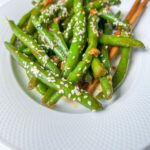 A white plate of Asian style green beans sautéed and garnished with sesame seeds, with wooden chopsticks resting on the side.