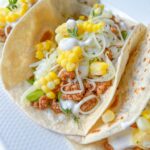 Three soft Ground Beef Tacos filled with seasoned meat, shredded cheese, corn kernels, lettuce, and a drizzle of white sauce, served on a white plate.
