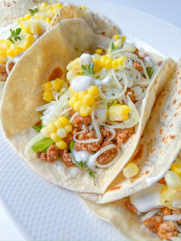 Three soft Ground Beef Tacos filled with seasoned meat, shredded cheese, corn kernels, lettuce, and a drizzle of white sauce, served on a white plate.