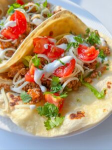 Two soft Ground Beef Tacos filled with seasoned meat, shredded cheese, lettuce, diced tomatoes, fresh cilantro, and drizzled with white sauce, served on a white plate.