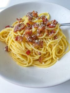 A white bowl filled with classic carbonara spaghetti, topped with crispy bacon pieces and grated cheese, with a fork resting on the side.