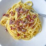 A plate of classic Carbonara topped with crispy pancetta and grated cheese, served on a white dish with a fork.