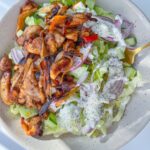 A bowl of Chicken Fajita Salad topped with grilled chicken, sliced red onions, lettuce, cucumber, and drizzled with a creamy dressing, served with a gold fork.