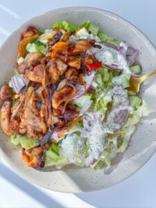 A bowl of Chicken Fajita Salad topped with grilled chicken, sliced red onions, lettuce, cucumber, and drizzled with a creamy dressing, served with a gold fork.