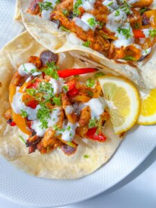 Two soft tortillas topped with grilled chicken, red and yellow bell peppers, fresh herbs, and a drizzle of white sauce create these Easy Chicken Fajitas, served with lemon wedges on a white plate.