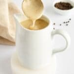 A white ceramic pitcher filled with creamy pepper gravy, with a spoon lifting some out. In the background are a beige cloth and a small bowl of black peppercorns—perfect for serving this easy gravy recipe on any occasion.