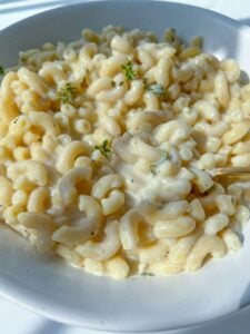 A close-up of creamy Mac and Cheese in a white bowl, topped with a few sprigs of fresh herbs, with a fork partially visible mixing the pasta.