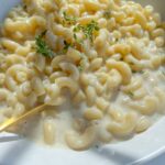 A bowl of creamy Mac and Cheese garnished with fresh sprigs of thyme, with a gold fork resting in the dish. The smooth, glossy sauce coats every elbow macaroni for the ultimate comfort food experience.