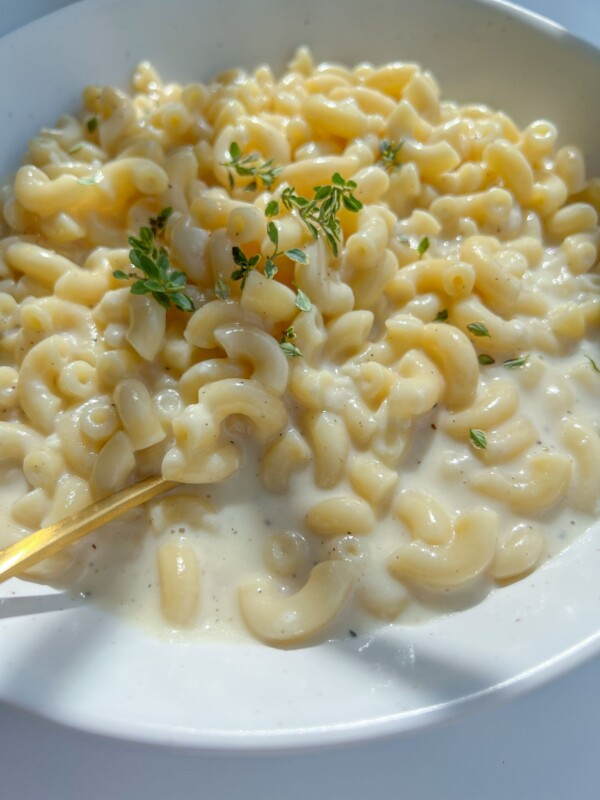 A bowl of creamy Mac and Cheese garnished with fresh sprigs of thyme, with a gold fork resting in the dish. The smooth, glossy sauce coats every elbow macaroni for the ultimate comfort food experience.