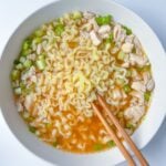 A bowl of ramen noodle soup with pieces of chicken and chopped green onions, served with a pair of wooden chopsticks on a white surface, makes for a comforting and flavorful ramen noodle soup experience.