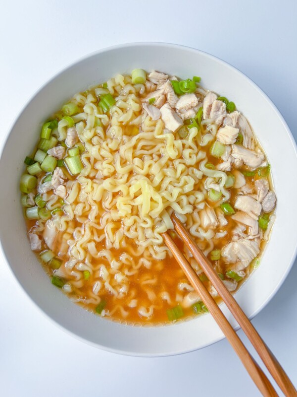 A bowl of ramen noodle soup with pieces of chicken and chopped green onions, served with a pair of wooden chopsticks on a white surface, makes for a comforting and flavorful ramen noodle soup experience.