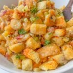 A bowl of homemade stuffing made with golden toasted bread cubes, diced celery, herbs, and small bits of bacon, garnished with fresh thyme—this is truly the Best Stuffing Recipe, with a wooden spoon partially visible.