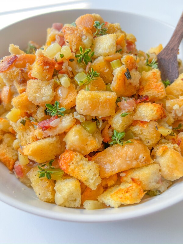 A bowl of homemade stuffing made with golden toasted bread cubes, diced celery, herbs, and small bits of bacon, garnished with fresh thyme—this is truly the Best Stuffing Recipe, with a wooden spoon partially visible.