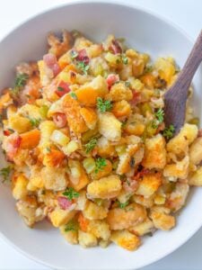A bowl of the Best Stuffing Recipe featuring golden toasted bread cubes, diced celery, bacon bits, and fresh herbs, with a wooden spoon resting in the dish.